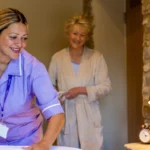 A carer in a purple uniform makes a bed while an older woman in a dressing gown smiles and watches in a cosy, warmly lit bedroom with stone walls and a lamp on a bedside table.