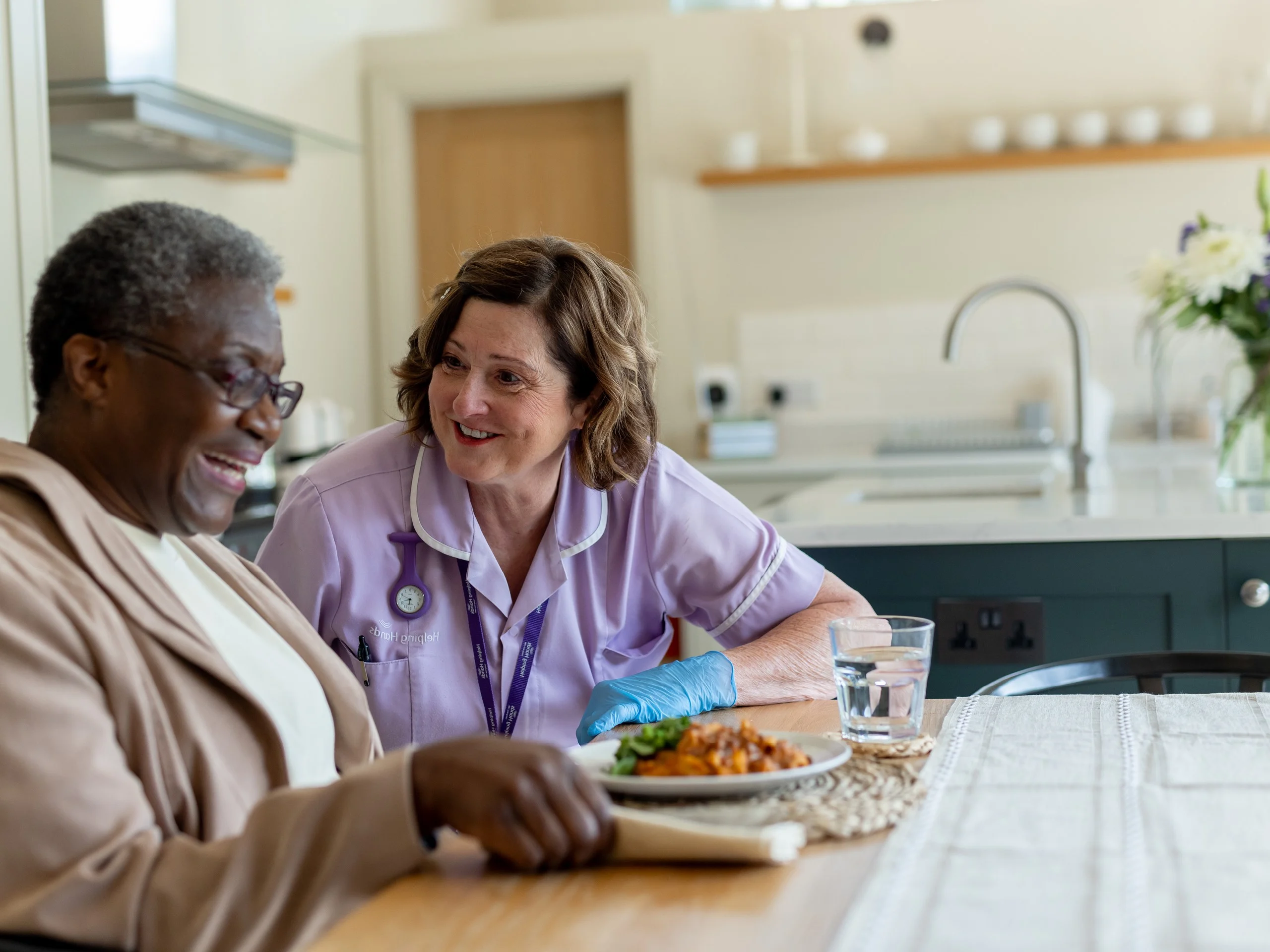 A smiling elderly adult sits at a table with a meal, while a carer in a purple uniform and gloves sits beside them, engaged in friendly conversation in a bright kitchen.