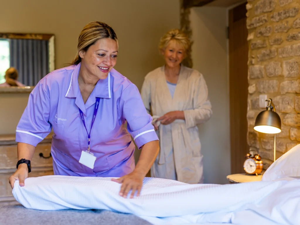 A smiling carer in a purple uniform makes a bed while an older woman in a dressing gown stands nearby, smiling, in a cosy, well-lit bedroom with stone walls and a bedside lamp.