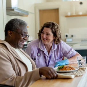 A smiling carer in a purple uniform and gloves sits next to an older woman who is laughing and eating a meal at a kitchen table. A glass of water and a plate of food are on the table.