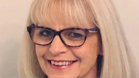 Smiling woman with straight, light blonde hair and fringe, wearing dark-framed glasses and a light-coloured top, pictured against a plain, light background.