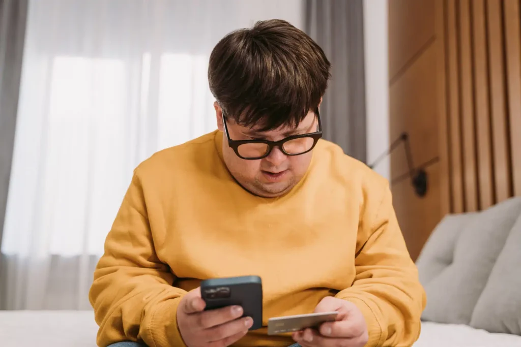 A person with Down's syndrome wearing glasses and a yellow sweatshirt sits on a bed, looking at a mobile phone in one hand and holding a credit card in the other.