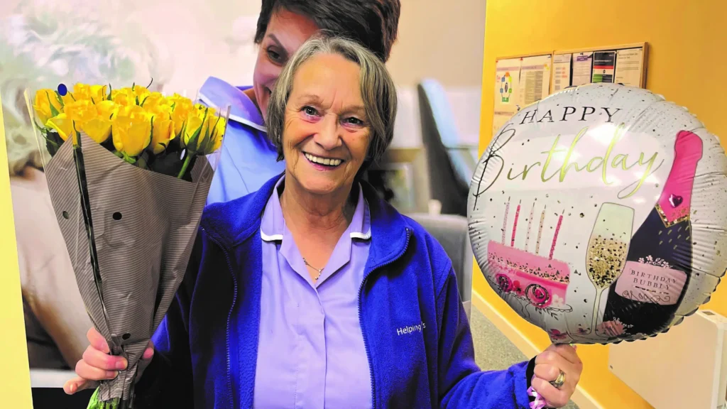 Smiling elderly woman in a blue jacket holds a bouquet of yellow roses in one hand and a Happy Birthday balloon in the other, with another person in the background.