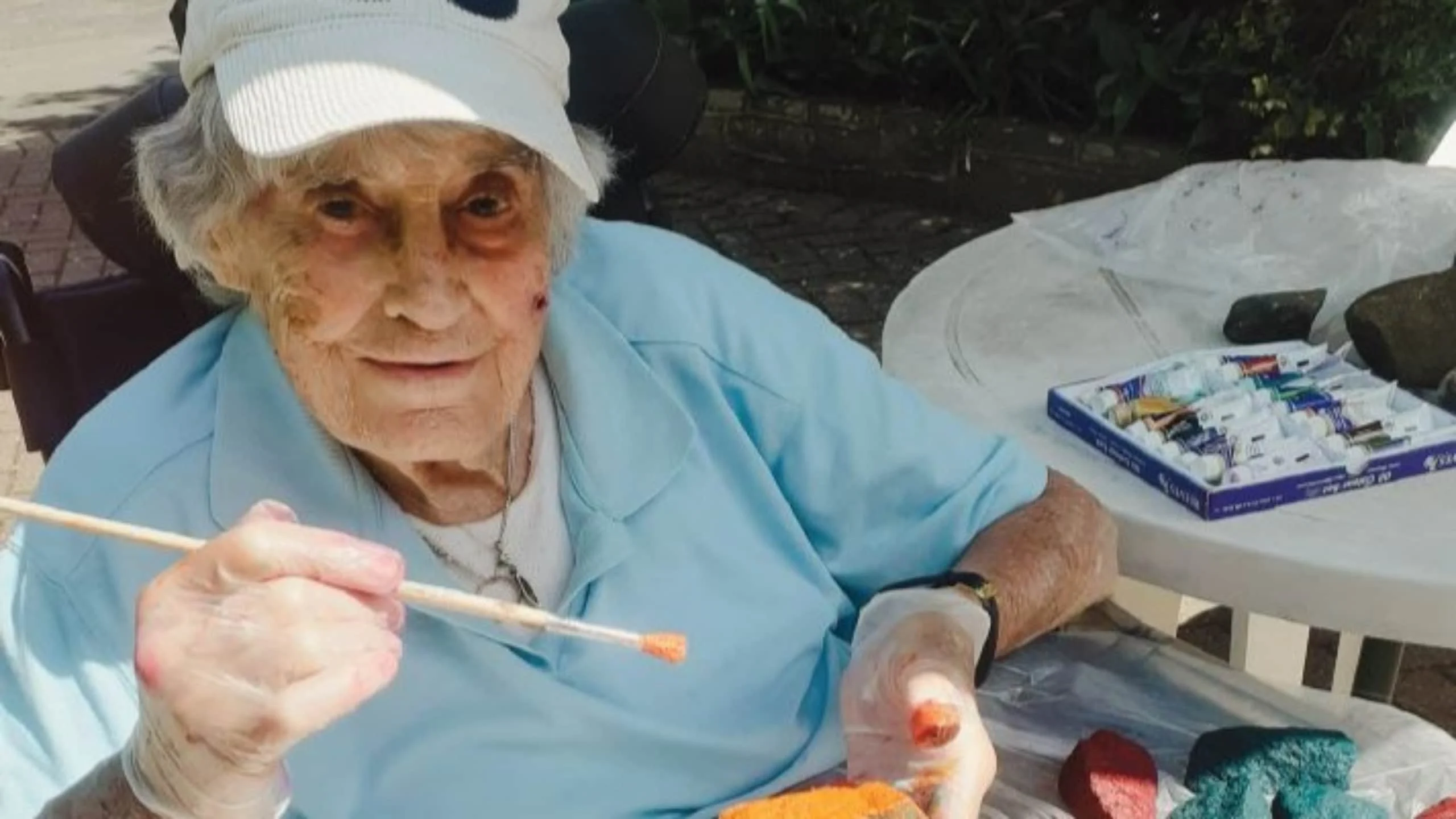 An elderly person wearing a white cap and blue shirt sits at a table outdoors, holding a paintbrush and painting a rock orange. Art supplies and rocks are on the table nearby.