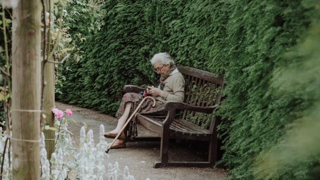 elderly woman sitting on bench by green hedge 16x9..