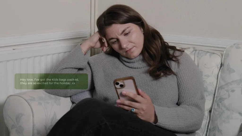A woman with long brown hair, wearing a grey jumper and black trousers, sits on a sofa looking at her phone with a concerned expression. A message on her screen reads: Hey love, I’ve got the kids’ bags packed, they are so excited for the holiday. xx.