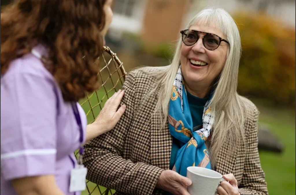 An older woman with long grey hair, sunglasses, and a patterned scarf smiles whilst holding a cup. She sits outdoors, talking with a person in a purple uniform who touches her arm supportively.