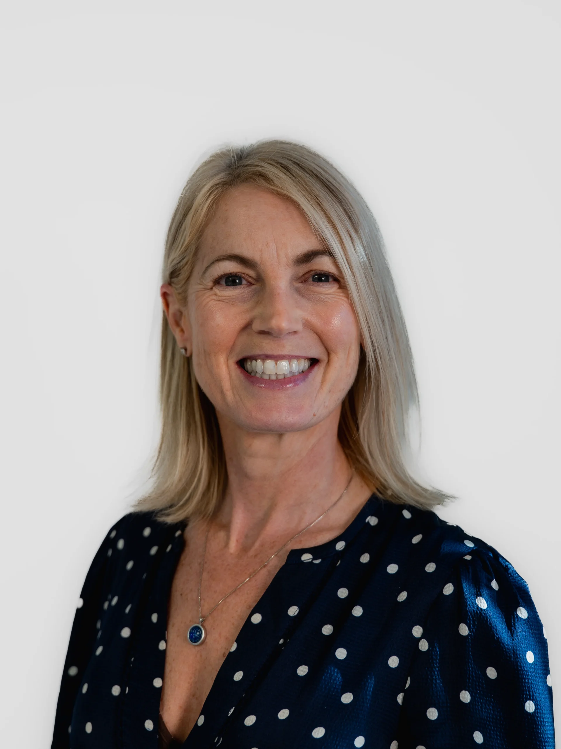 A smiling woman with straight blonde hair wearing a navy blue blouse with white polka dots and a necklace, standing against a plain light background.