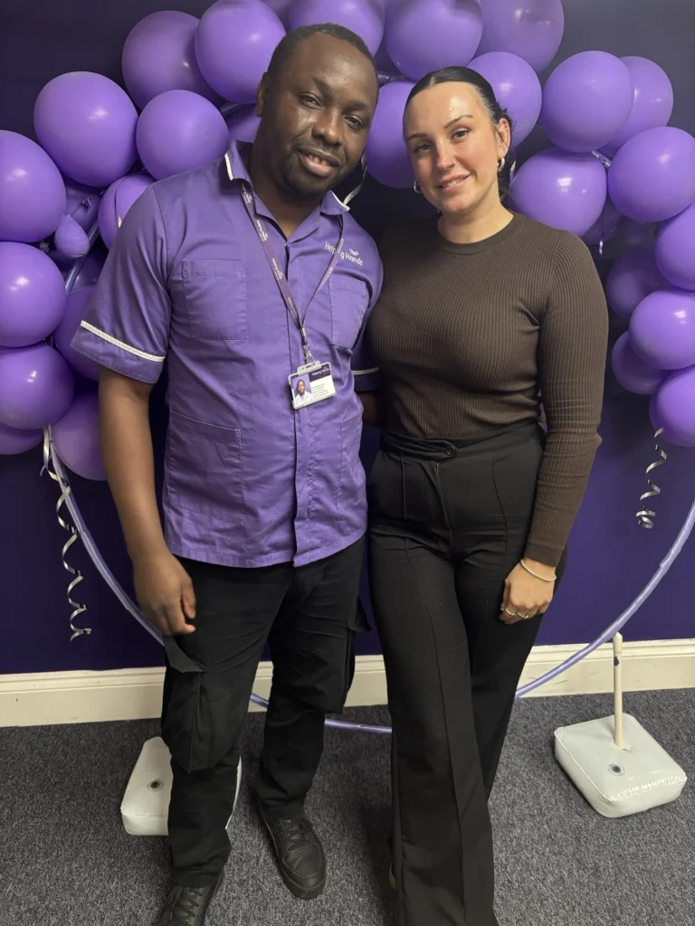 Two people standing and smiling in front of a purple wall decorated with clusters of purple balloons. One person wears a purple uniform with a name badge; the other wears a brown top and black trousers.