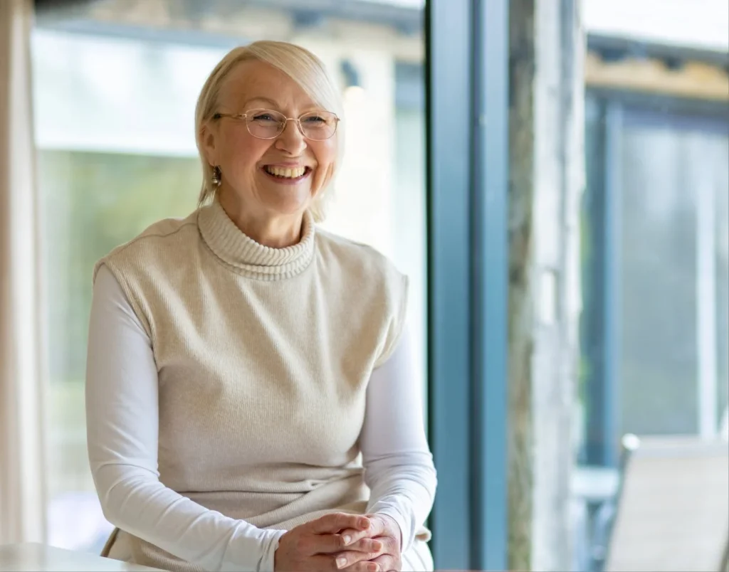 An older woman with glasses and blonde hair smiles whilst sitting indoors by a large window, wearing a white polo neck and a beige sleeveless jumper. Natural light fills the modern, bright space.