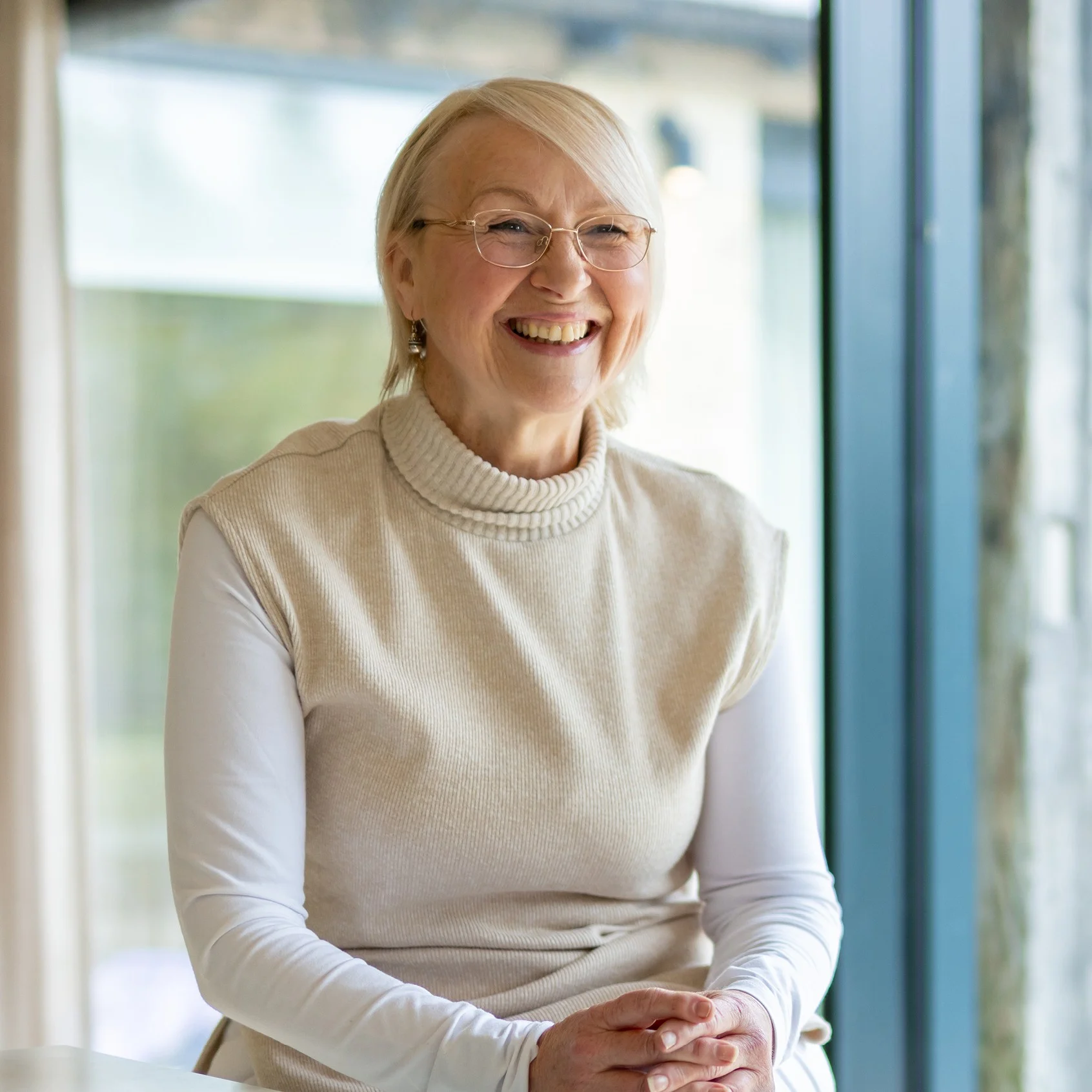 An older woman with glasses and short blonde hair smiles whilst sitting by a large window, wearing a light beige sleeveless jumper over a white long-sleeve shirt.