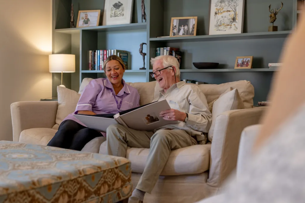 An elderly man and a woman sit smiling on a beige sofa, looking at a large photo album together in a cosy living room with shelves, books, and framed pictures in the background.