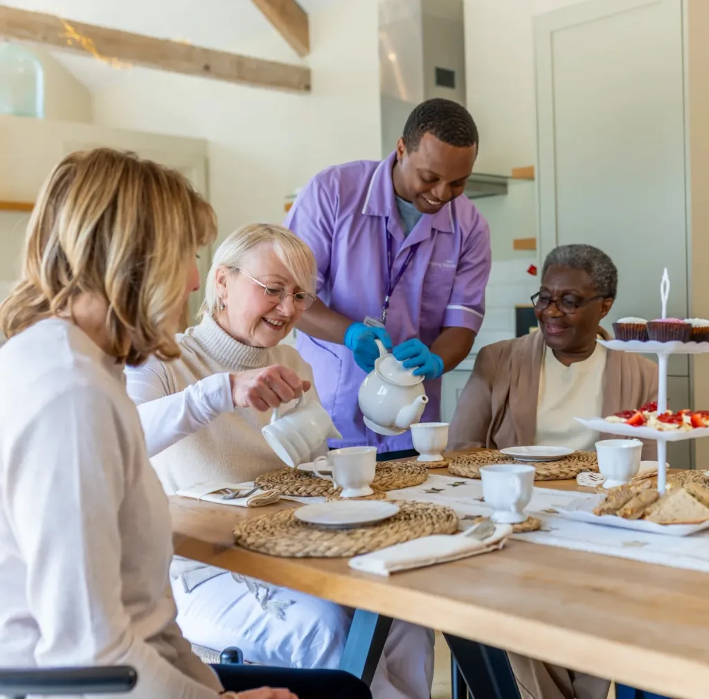 Three older women sit at a table set with tea and treats. A carer in purple scrubs and blue gloves helps serve tea. The group appears to be enjoying a friendly, relaxed gathering in a bright, cosy kitchen.