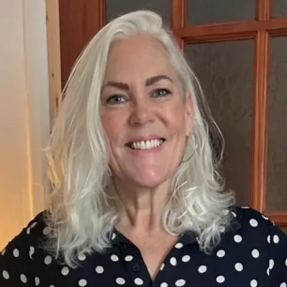 A woman with shoulder-length white hair smiles at the camera. She is wearing a navy blue shirt with white polka dots and standing in front of a wooden door with glass panels.
