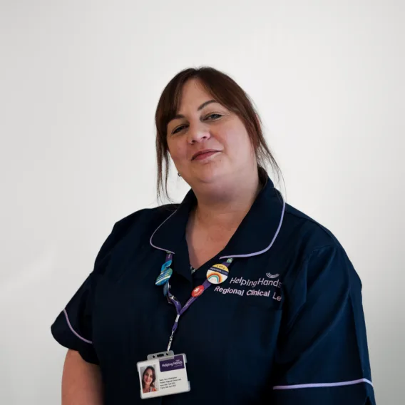 A woman in a navy blue uniform with purple trim stands against a plain white background, wearing an ID badge and lanyard. She smiles slightly and has brown hair tied back. The uniform reads Helping Hands regional clinical..
