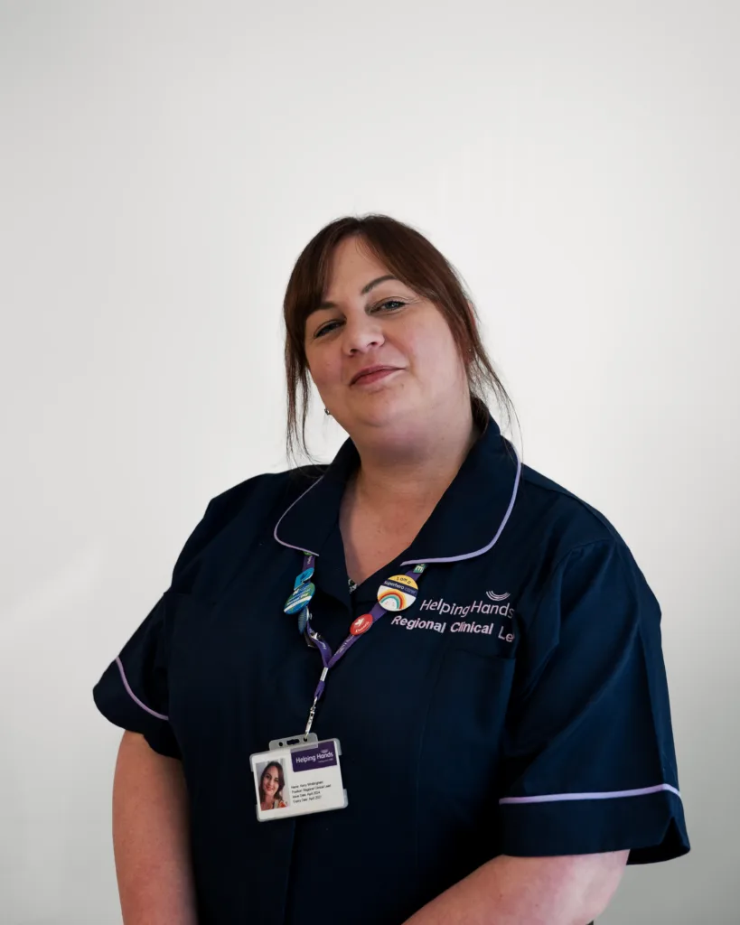 A woman in a navy blue uniform with purple trim stands against a plain white background, wearing an ID badge and lanyard. She smiles slightly and has brown hair tied back. The uniform reads Helping Hands regional clinical..