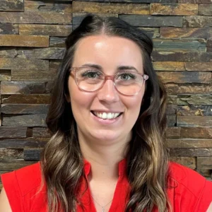 Smiling woman with long brown hair wearing glasses and a red top, standing in front of a textured stone wall.