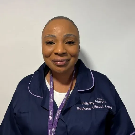 A person smiles at the camera, wearing a navy blue uniform with white piping and a purple lanyard. The uniform reads Helping Hands Regional Clinical Lead. The background is plain white.
