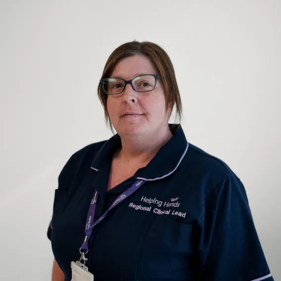 A woman wearing glasses and a dark blue Helping Hands Regional Clinical Lead uniform stands against a plain white background, facing the camera.