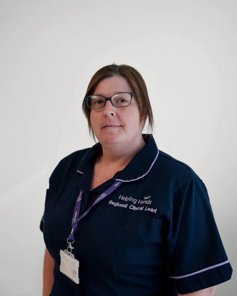 A woman wearing glasses and a dark blue Helping Hands Regional Clinical Lead uniform stands against a plain white background, facing the camera.