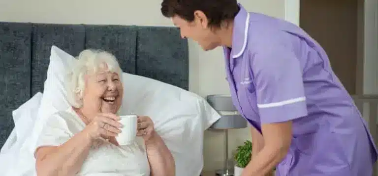 Elderly lady in bed with hot drink female carer purple uniform
