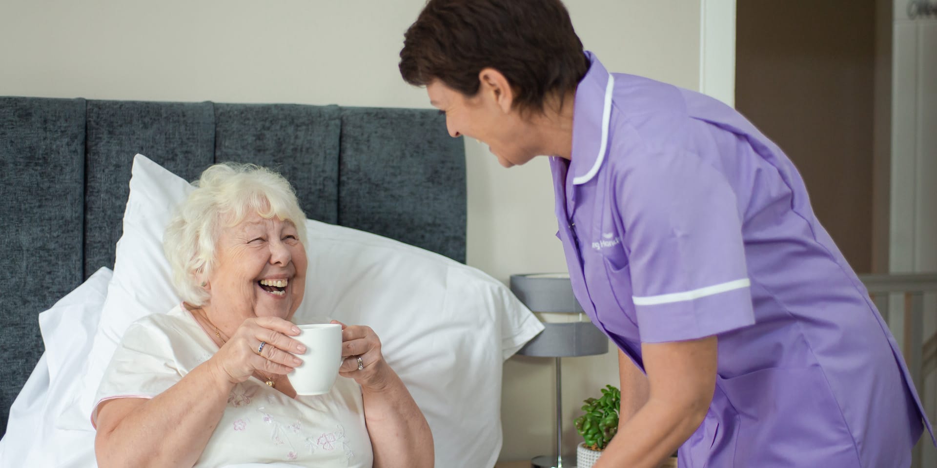 Elderly lady in bed with hot drink female carer purple uniform