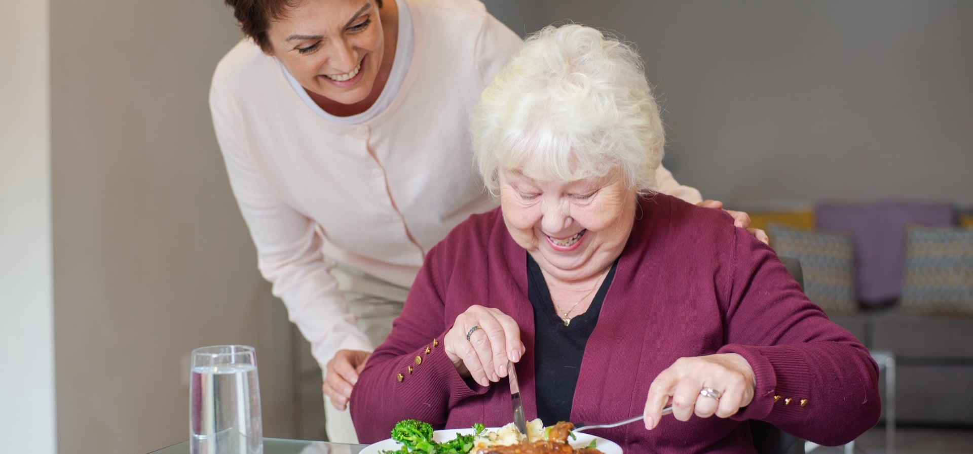 Elderly lady red cardigan eating using knife fork glass table female carer standing behind
