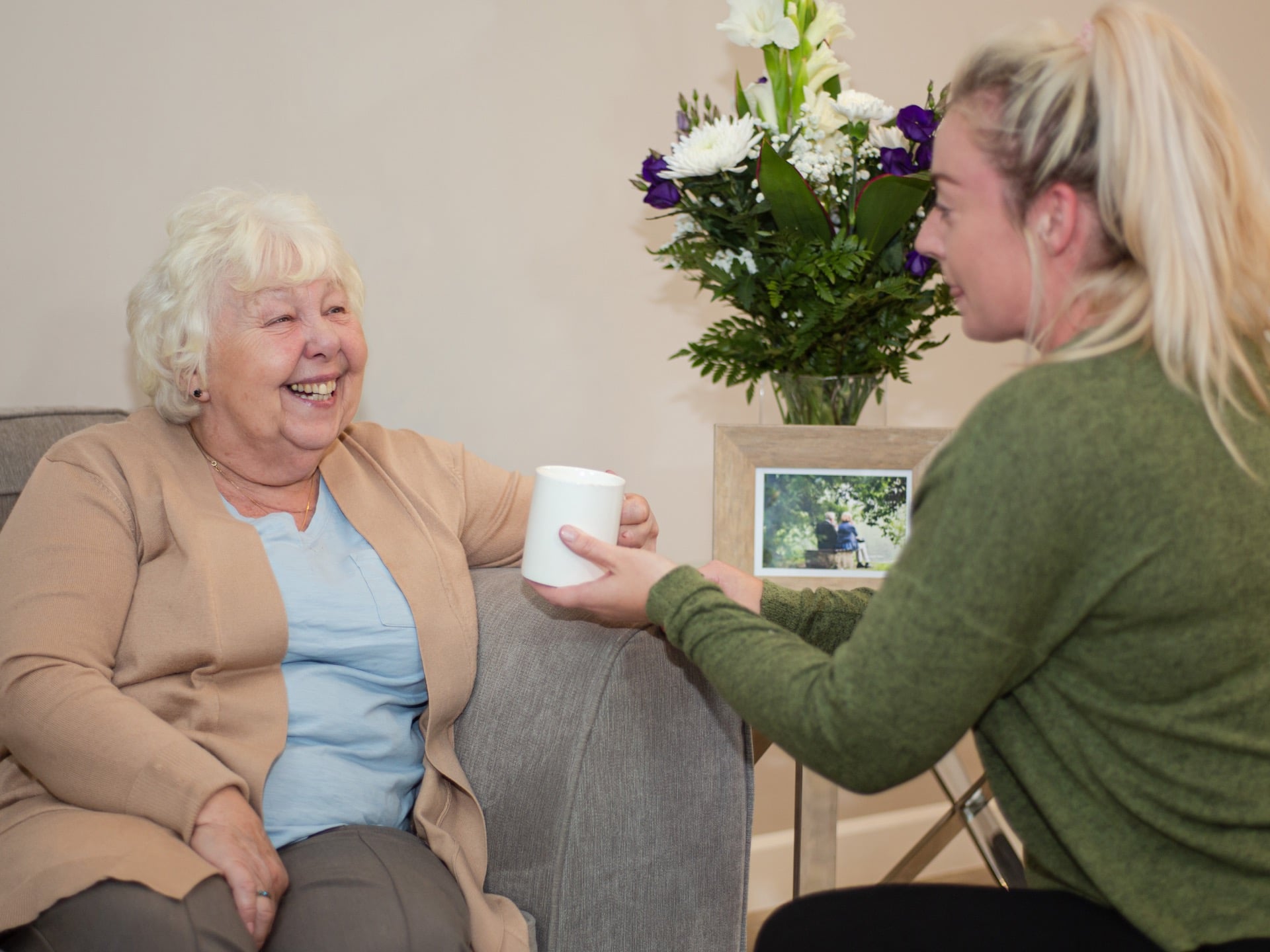 elderly-lady-sitting-on-sofa-female-carer-passing-mug_4x3