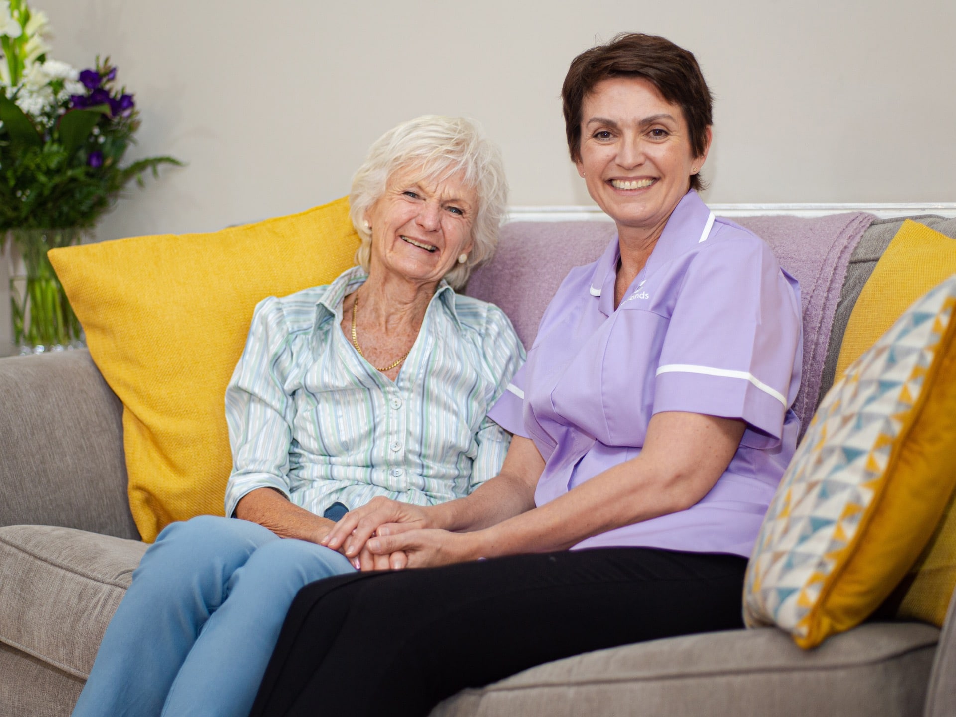 Elderly lady sitting on sofa with female carer holding hands smiling