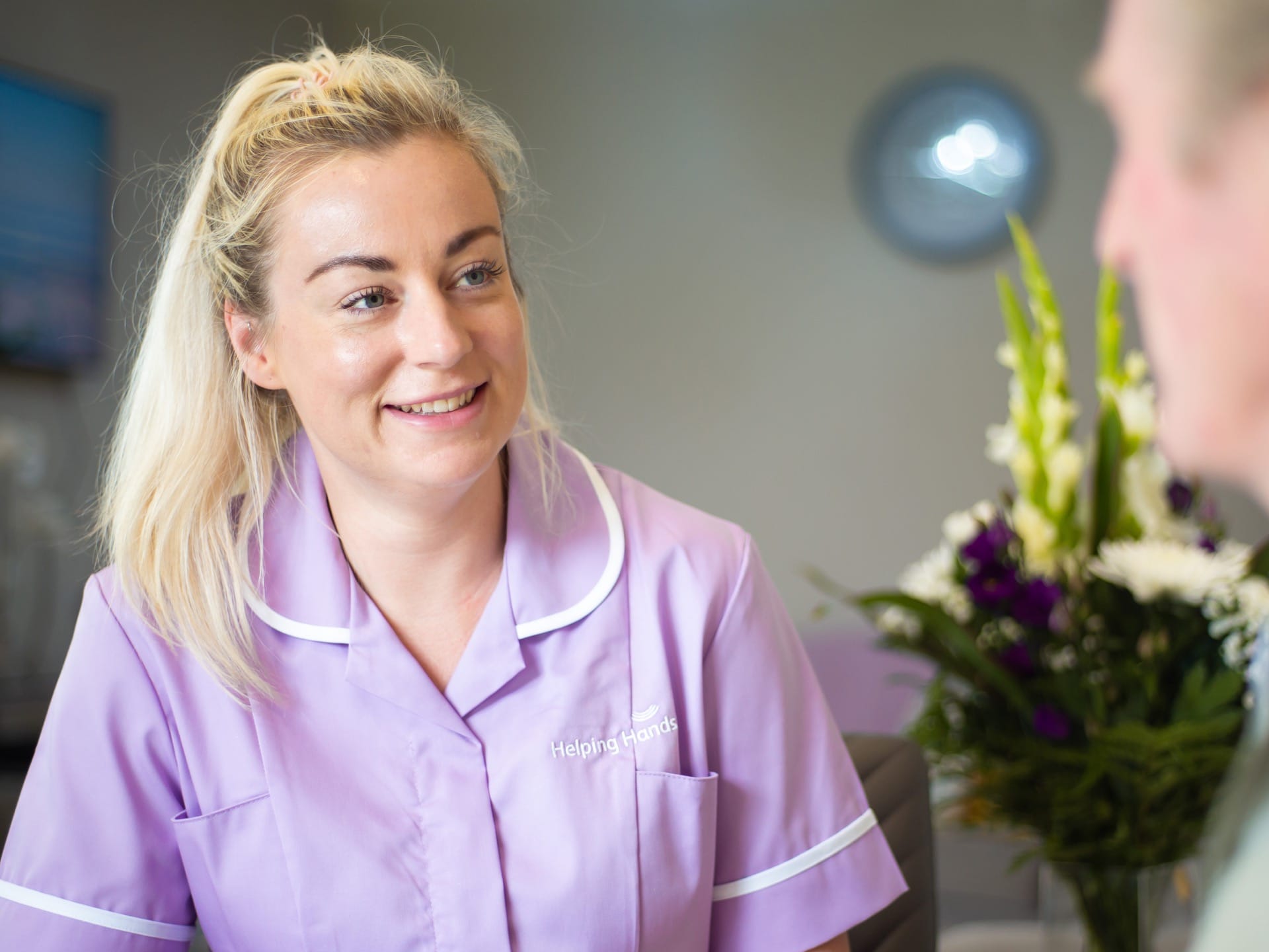 Female carer smiling purple uniform sitting with elderly male holding mug flowers