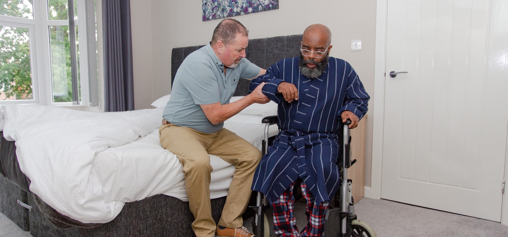 Male carer green polo top helping man in wheelchair holding arm blue gown