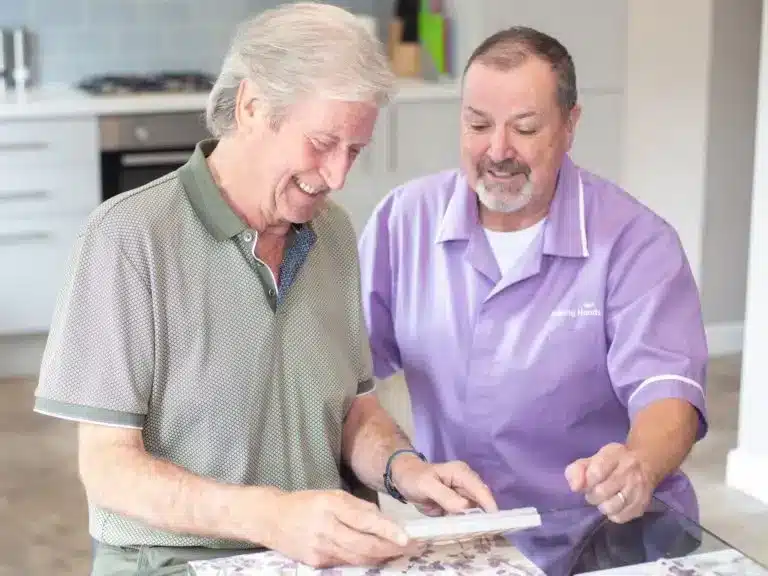 Male carer purple uniform crouching beside elderly man holding dosette box