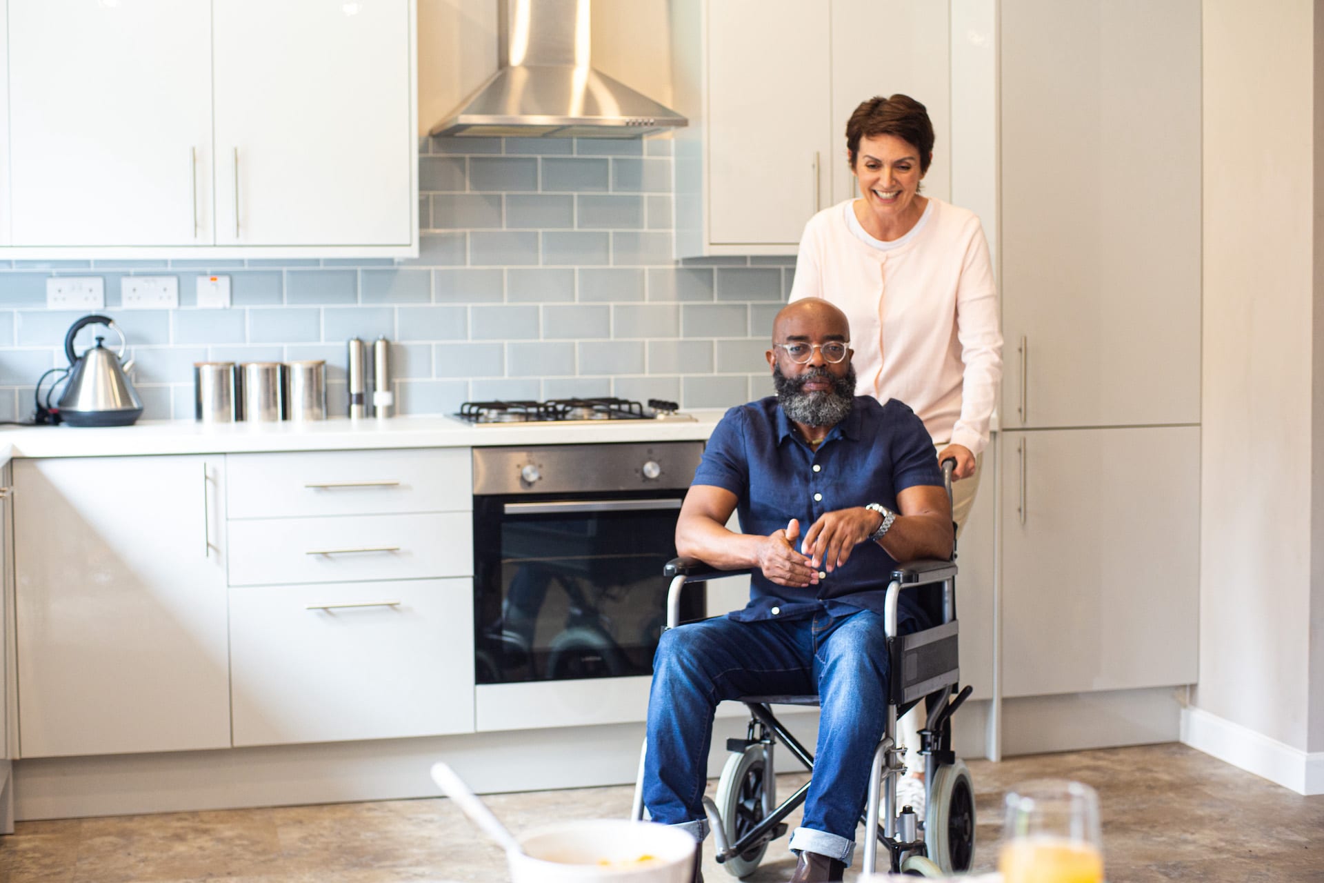 Man in wheelchair blue shirt female carer white top in kitchen